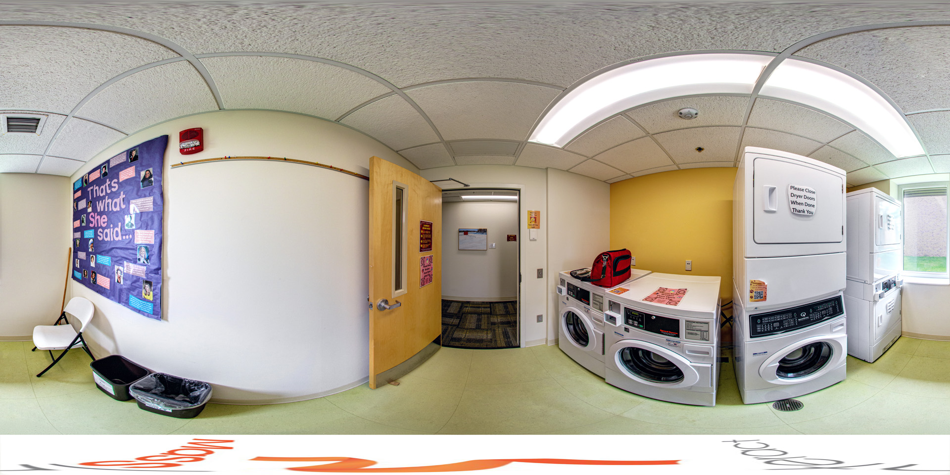 Panoramic view of a small laundry room with stacked washers and dryers, brightly lit by overhead lights. The space has a yellow accent wall and a window providing natural light. A folding chair and bins are placed next to a bulletin board on the opposite wall with a display titled, 'That's What She Said.'