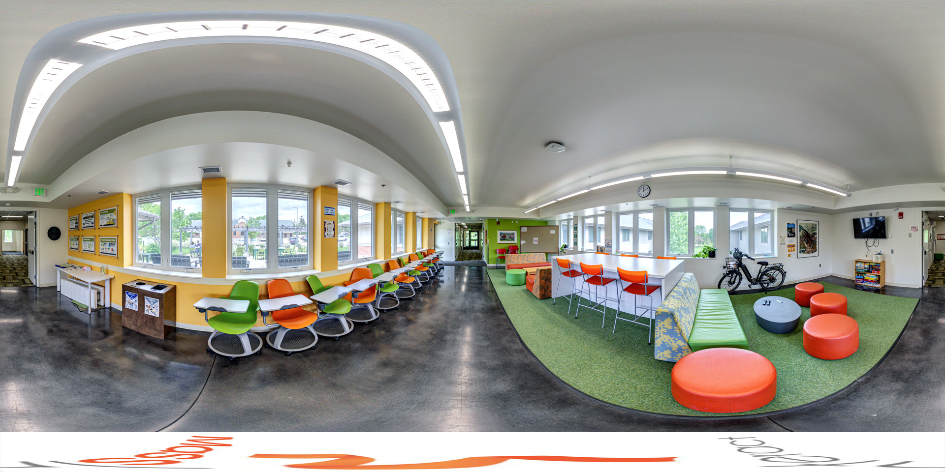 brightly lit lounge area with colorful seating options, including green and orange chairs, circular stools, and a long couch. The space has a large window, modern overhead lighting, and a green carpet in one section. A bicycle is parked near the wall, and a television is mounted above a shelf with board games.