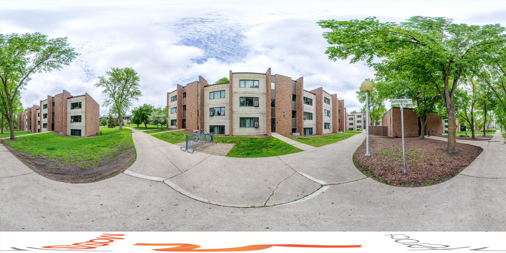 Panoramic view of of on-campus apartment buildings with a modern brick design, large windows, and balconies. A bike rack is near the entrance, surrounded by trees, grass, and sidewalks. A sign reads 'Switchgrass Ln.'' 