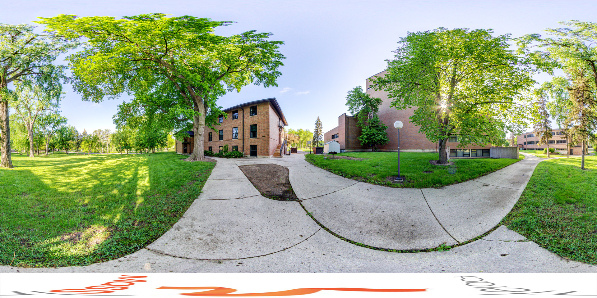 Panoramic view of a peaceful green campus area with large, leafy trees providing shade. The scene includes a brick building, sidewalks, and grassy areas, with sunlight filtering through the trees, casting long shadows. 