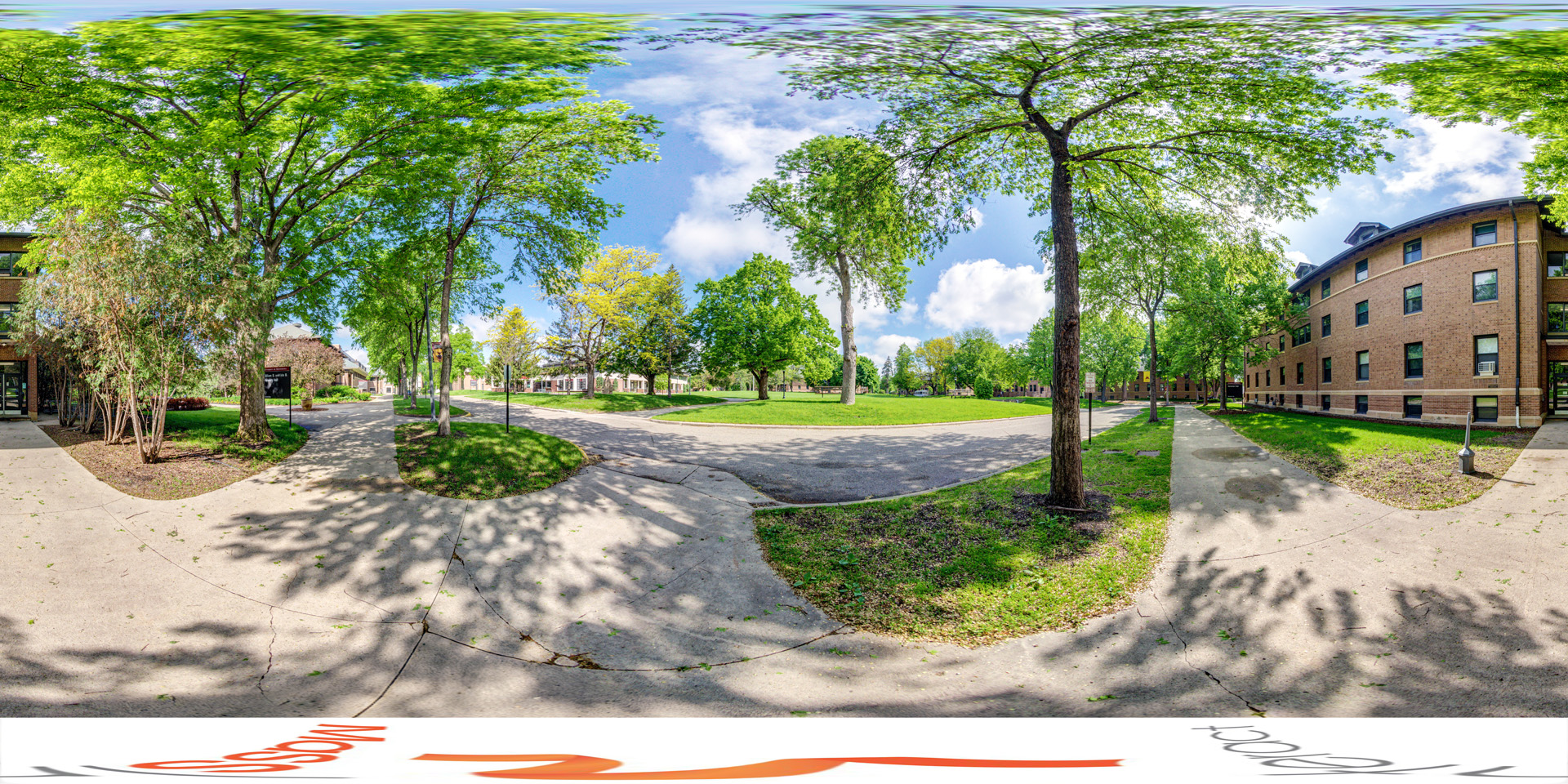 Panoramic view of a tranquil campus setting featuring a tree-lined pathway surrounded by green grass. The scene includes a brick building on the right, a clear sky, and a mix of leafy trees casting shadows on the walkways, creating a peaceful outdoor atmosphere.