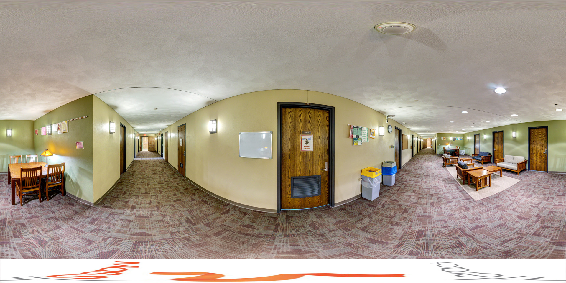 Panoramic view of a dormitory hallway with room doors on both sides, carpeted with a patterned design. To the left, there’s a small study area with a table, chairs, and a lamp. Further down, a lounge area features couches, chairs, and coffee tables. The space is well-lit with wall-mounted lights, and recycling bins are near the room entrances.