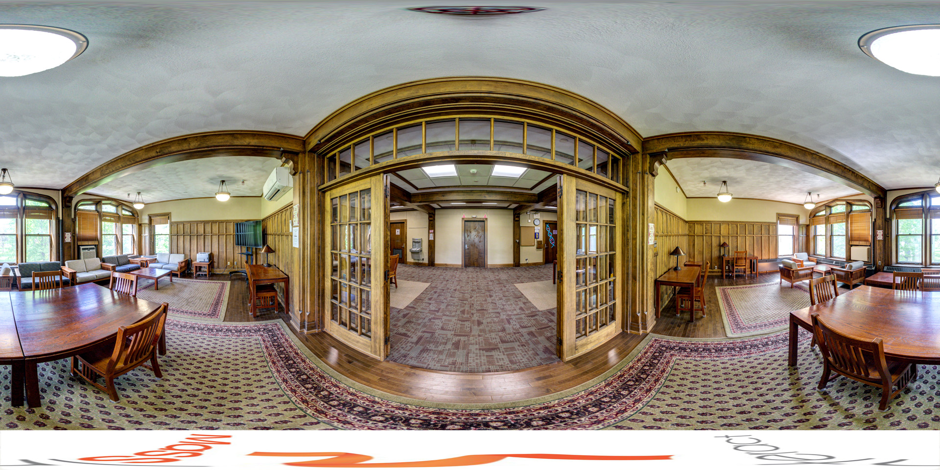 Panoramic view of a spacious lounge area with a vintage, wood-paneled design. The room is furnished with wooden tables, chairs, and soft seating arranged around patterned rugs. Large windows let in natural light, and a set of wooden, glass-paneled doors divides the space. The room has a cozy, classic atmosphere.