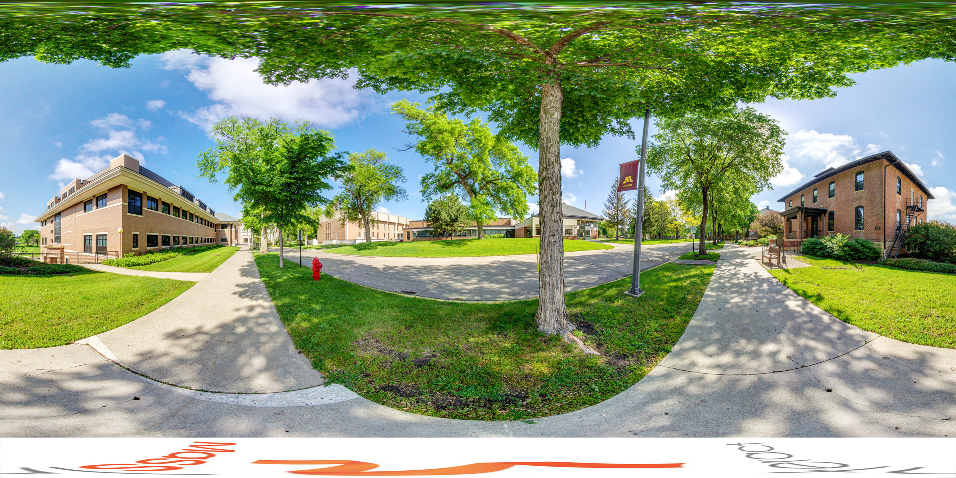 Panoramic view of a sunny campus scene with two brick buildings on either side of a paved walkway. The building on the left is modern, while the one on the right has a classic design with an outdoor staircase. Green trees provide shade, and a red fire hydrant is visible by the sidewalk. The open grassy area and clear blue sky create a peaceful atmosphere.