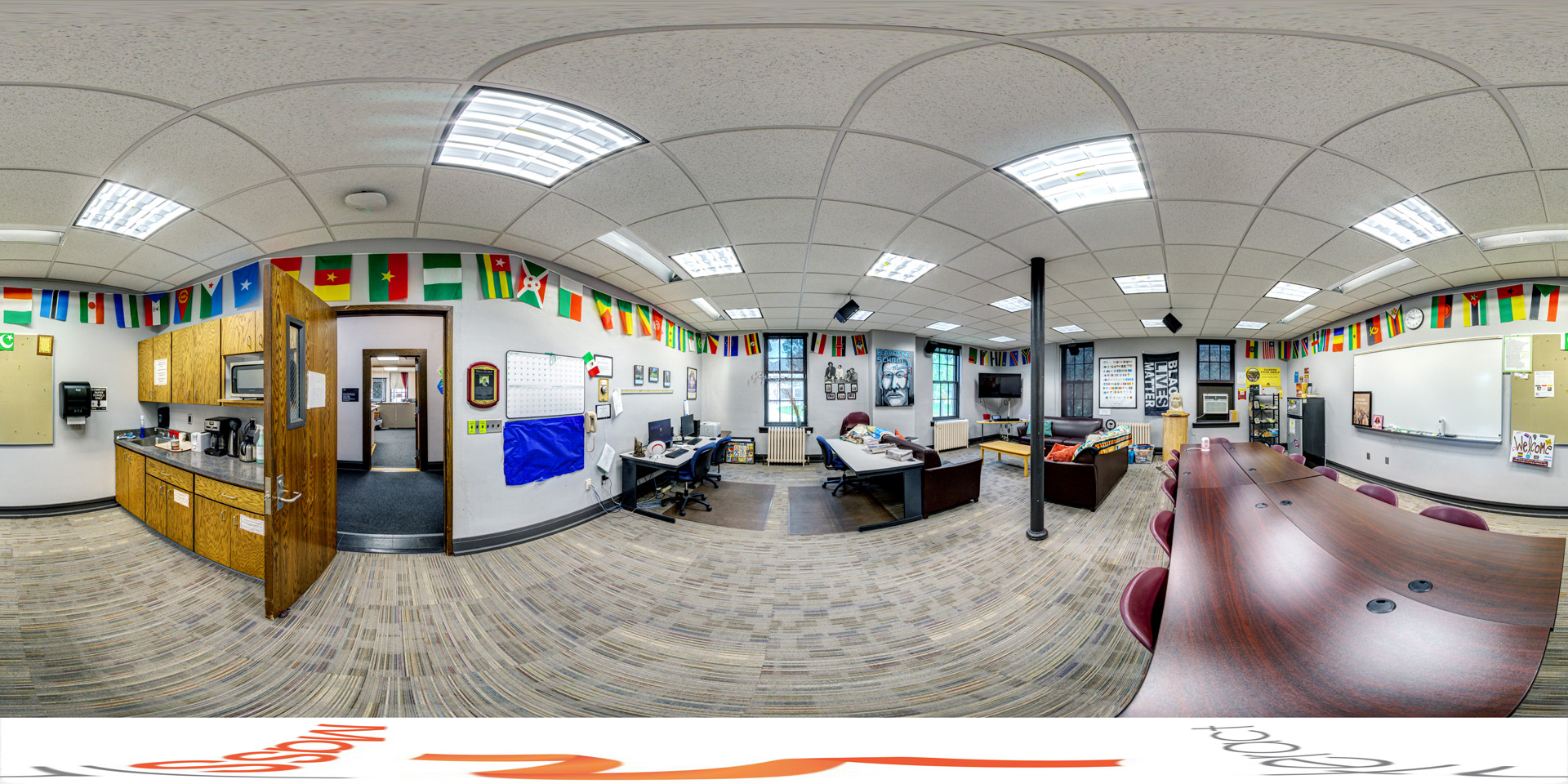 Panoramic view of a bright office lounge with a curved table and chairs, workstations with computers, and a cozy seating area. The walls are decorated with international flags, adding vibrant color. A small kitchenette includes a microwave and coffee makers. Posters and a 'Black Lives Matter' sign enhance the room's welcoming and inclusive atmosphere.