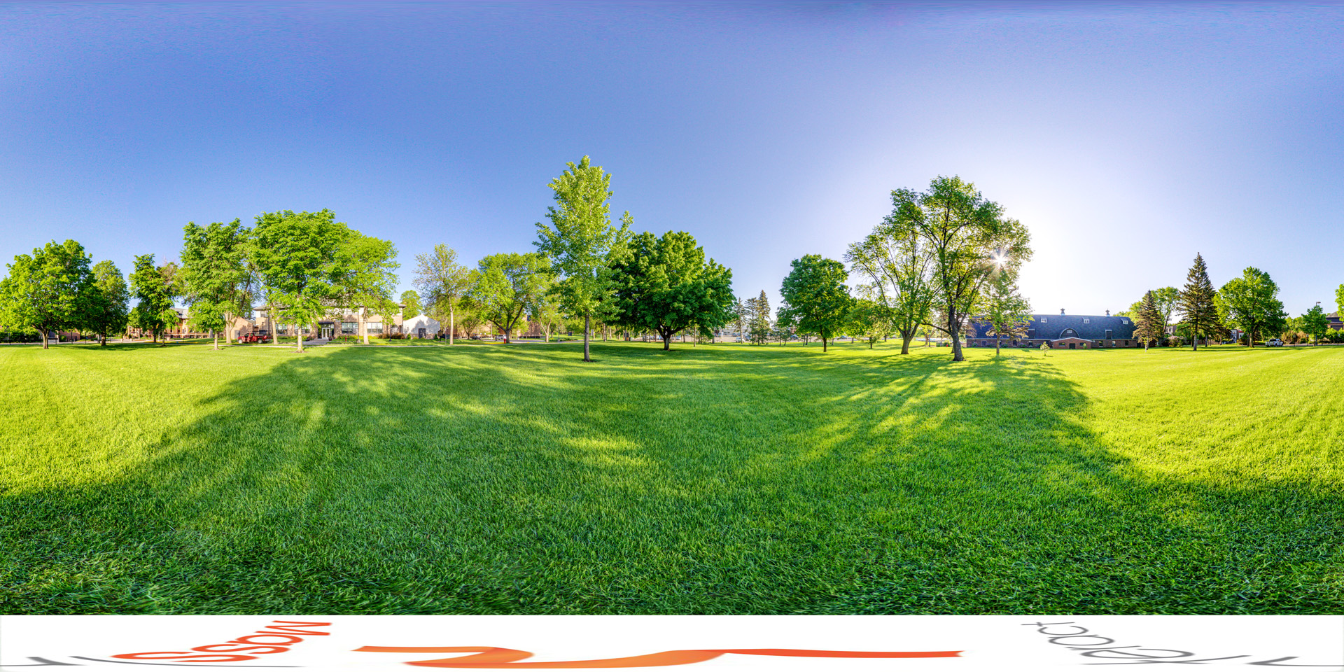 Panoramic view of a wide, grassy field on a sunny day, bordered by trees and buildings in the distance. The trees cast long shadows on the vibrant green grass, and the bright blue sky is clear and expansive. One of the visible structures in the background appears to be a large barn, suggesting a rural or agricultural setting.