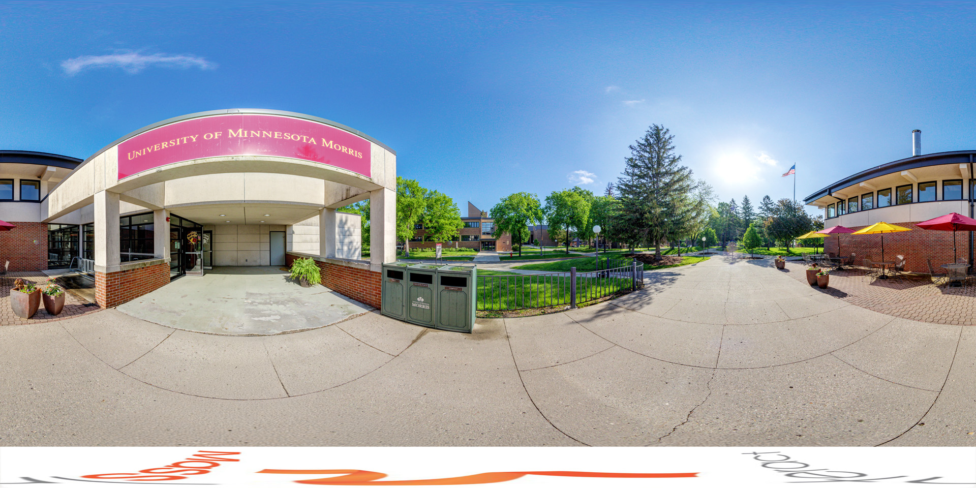 Panoramic view of a front view of the University of Minnesota Morris Student Center. The building features a covered entrance with a large sign displaying the university's name. To the right, there is an outdoor seating area with tables, chairs, and colorful umbrellas. In the distance, the campus is lined with trees, open green spaces, and an American flag flying on a pole.
