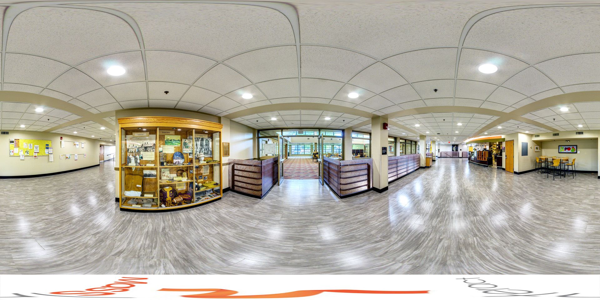 Panoramic view of a spacious hallway with light gray flooring and white ceiling tiles. To the right, there is a café area with bar-height tables and chairs. In the center, a large display case showcases memorabilia and historical items. Ahead, glass doors lead to a seating area with tables and chairs.