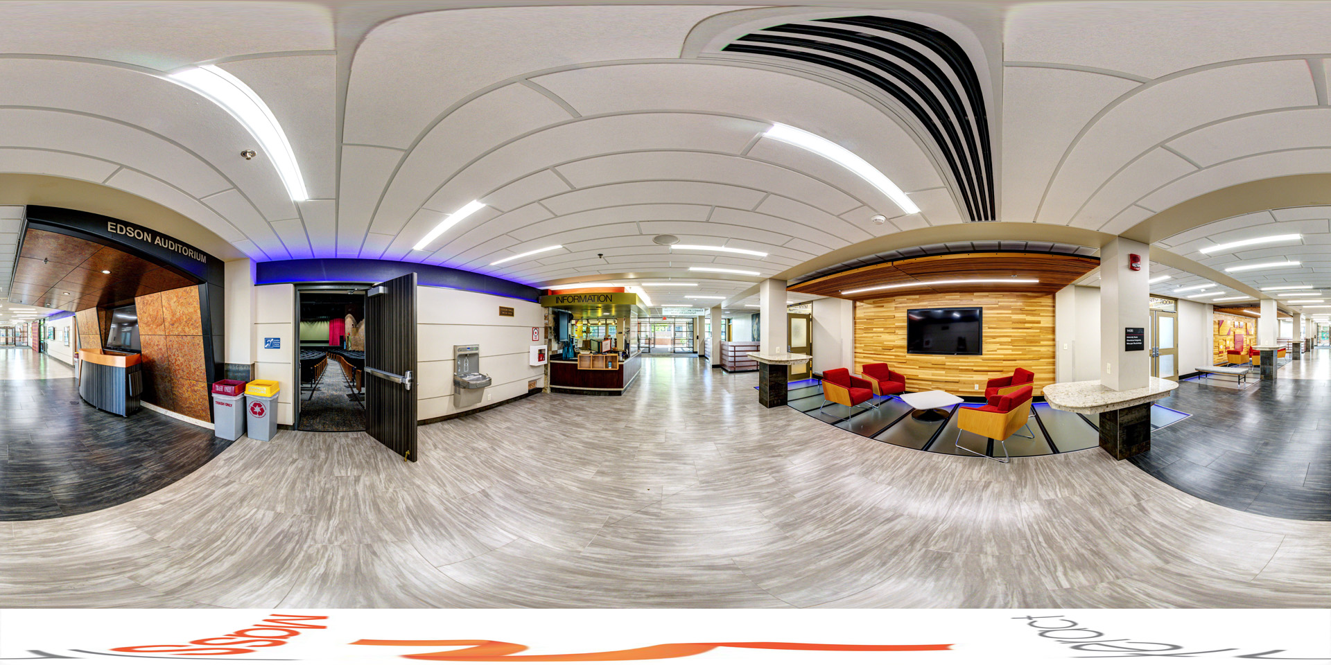 Panoramic view of a entrance to Edson Auditorium in the Edward J. and Helen Jane Morrison Performing Arts Center. The area features modern wood and tile flooring, recessed lighting, and a curved ceiling. A seating area with red chairs and a wall-mounted TV is to the right. The auditorium entrance is directly ahead, and an information desk is nearby.