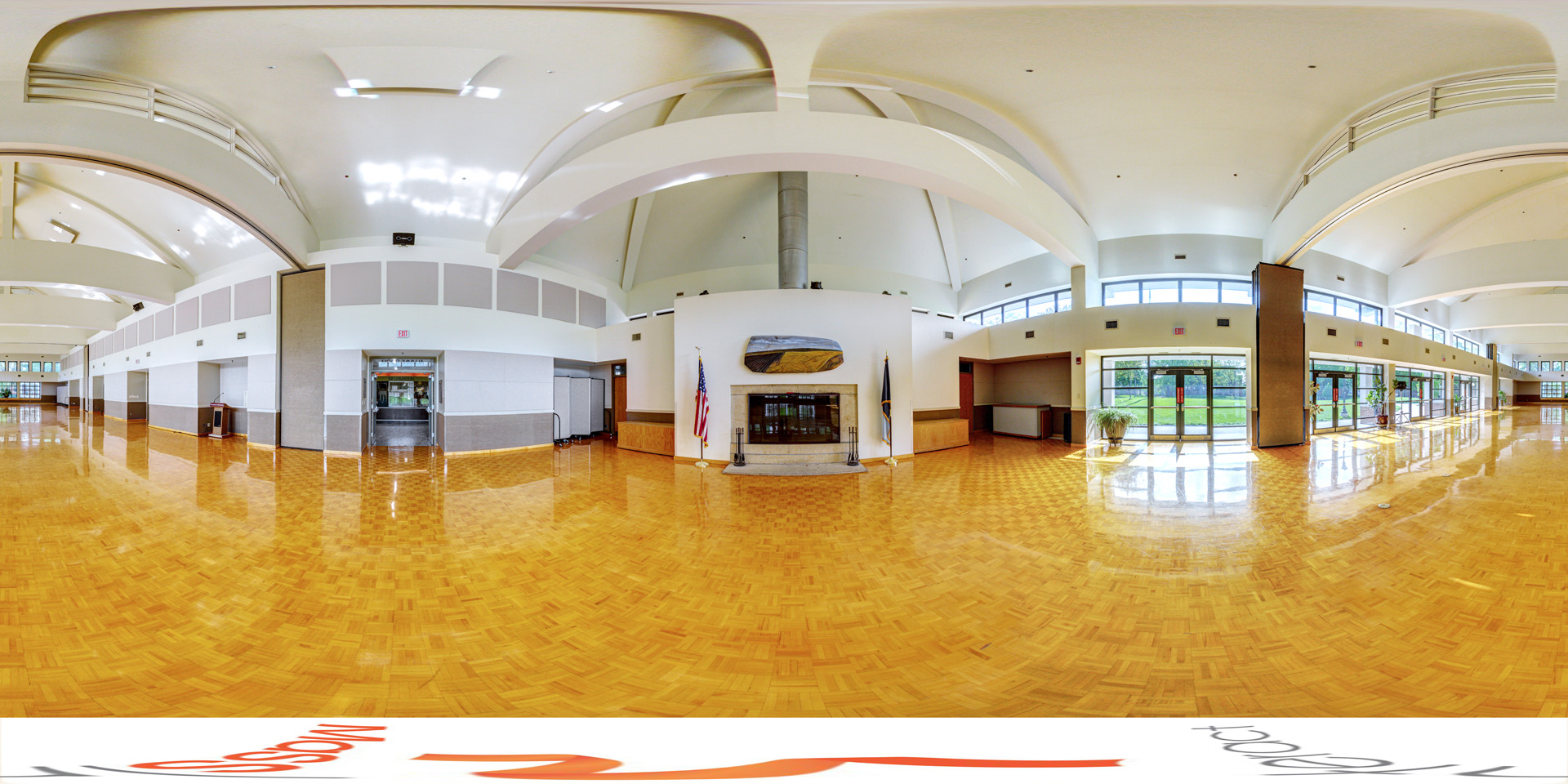 Panoramic view of a spacious hall with high, curved ceilings and polished wooden flooring. Large windows let in natural light, and there’s a central fireplace with flags on either side. The hall has multiple doors leading to outdoor areas, giving the space an open and bright atmosphere.