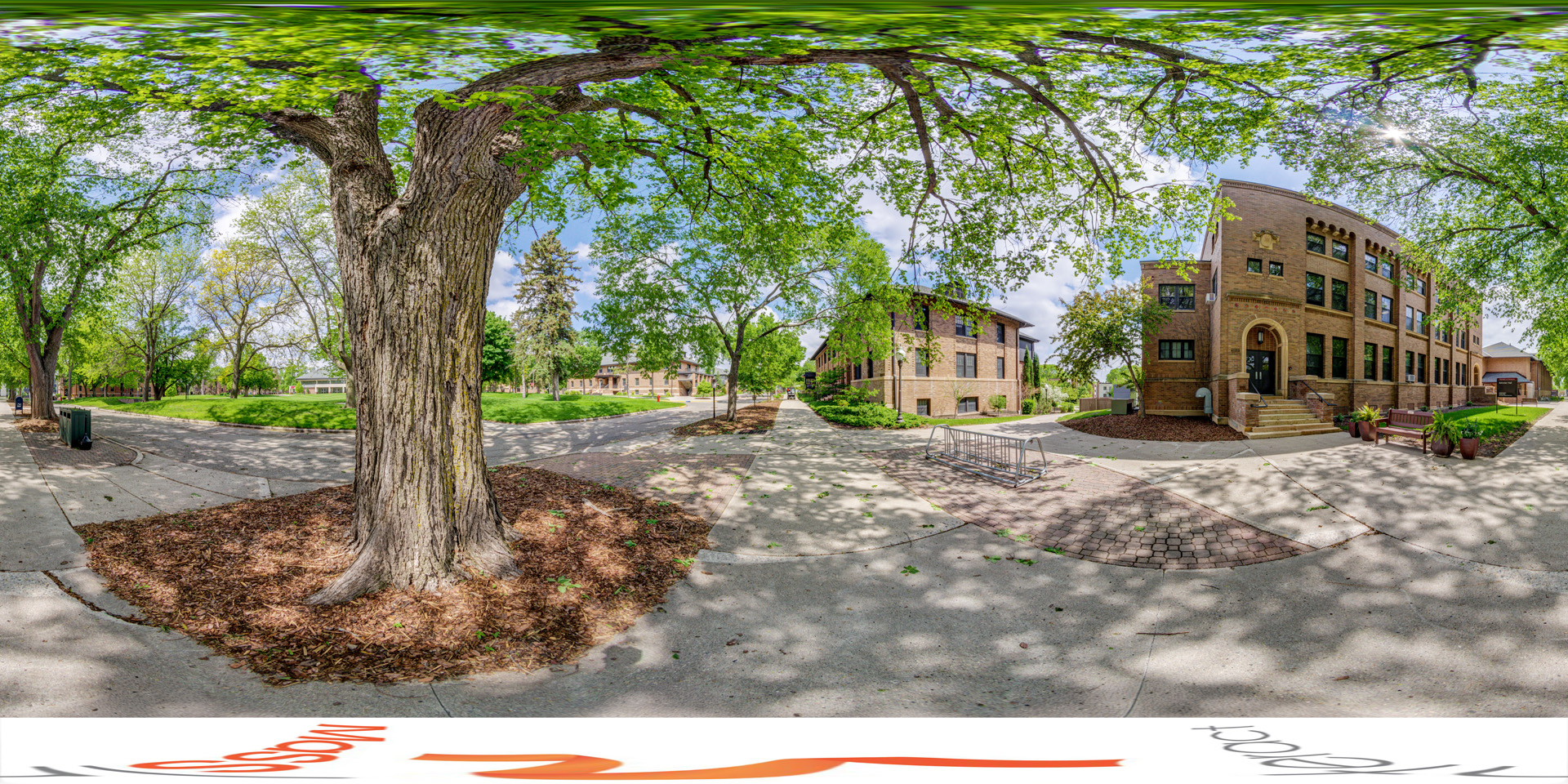 Panoramic view of a tree-lined sidewalk leading to a brick building with arched entryway, surrounded by shaded green spaces and other campus structures in the background.