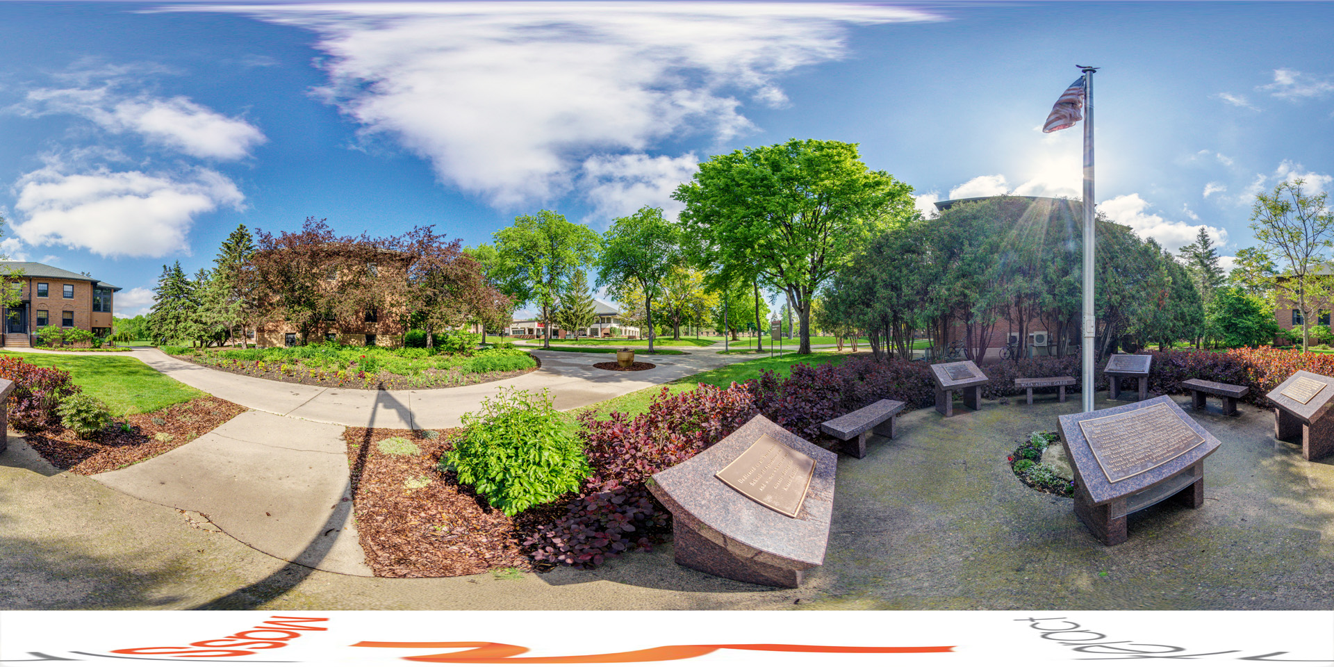 Panoramic view of a small circular memorial with stone plaques and benches arranged around a flagpole, surrounded by plants and trees on a well-maintained campus. A brick building is visible in the background, along with other campus buildings.