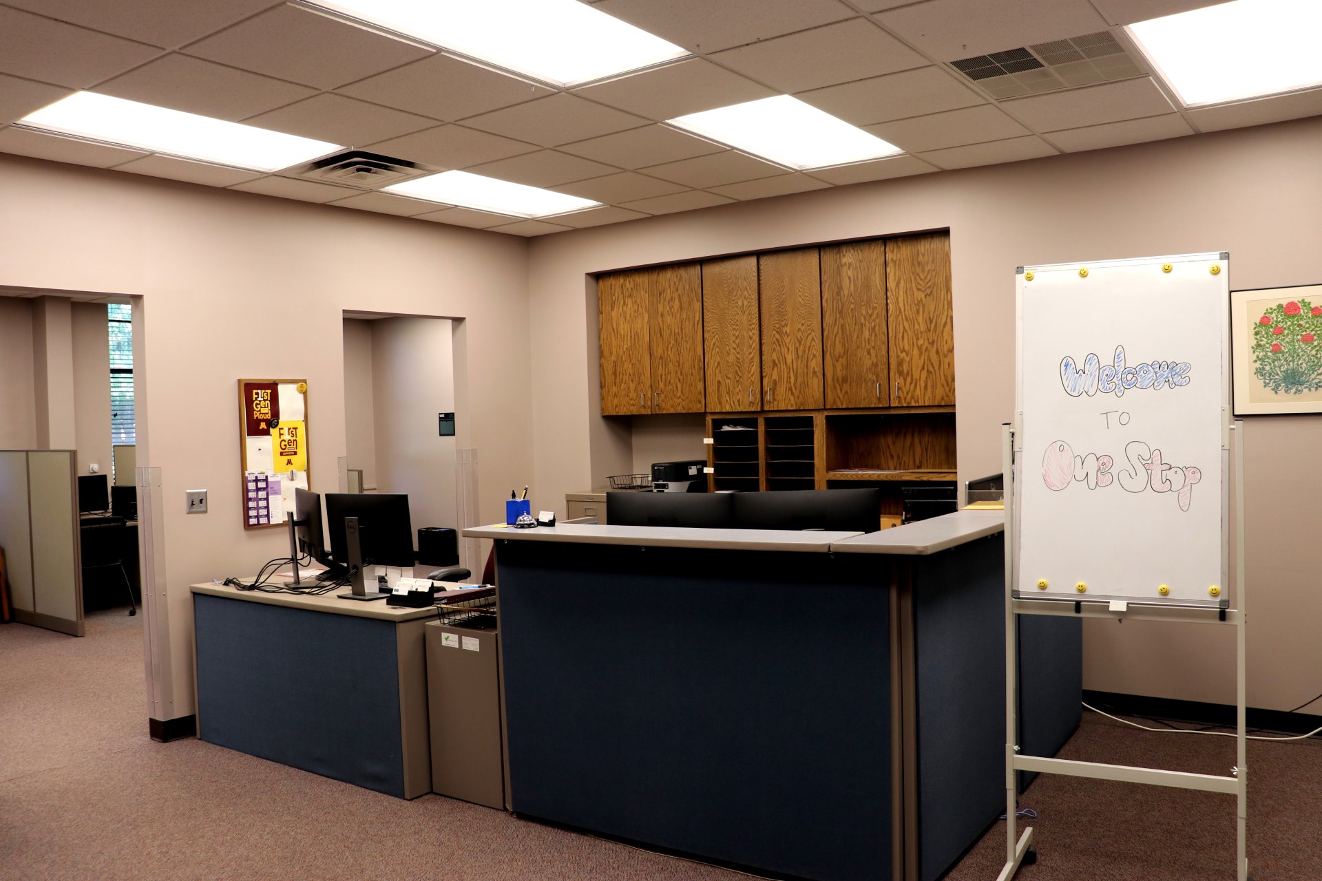 An office space with a central desk, computers, wooden cabinets, and a whiteboard that reads 'Welcome to One Stop.' The room is lit by overhead fluorescent lights.
