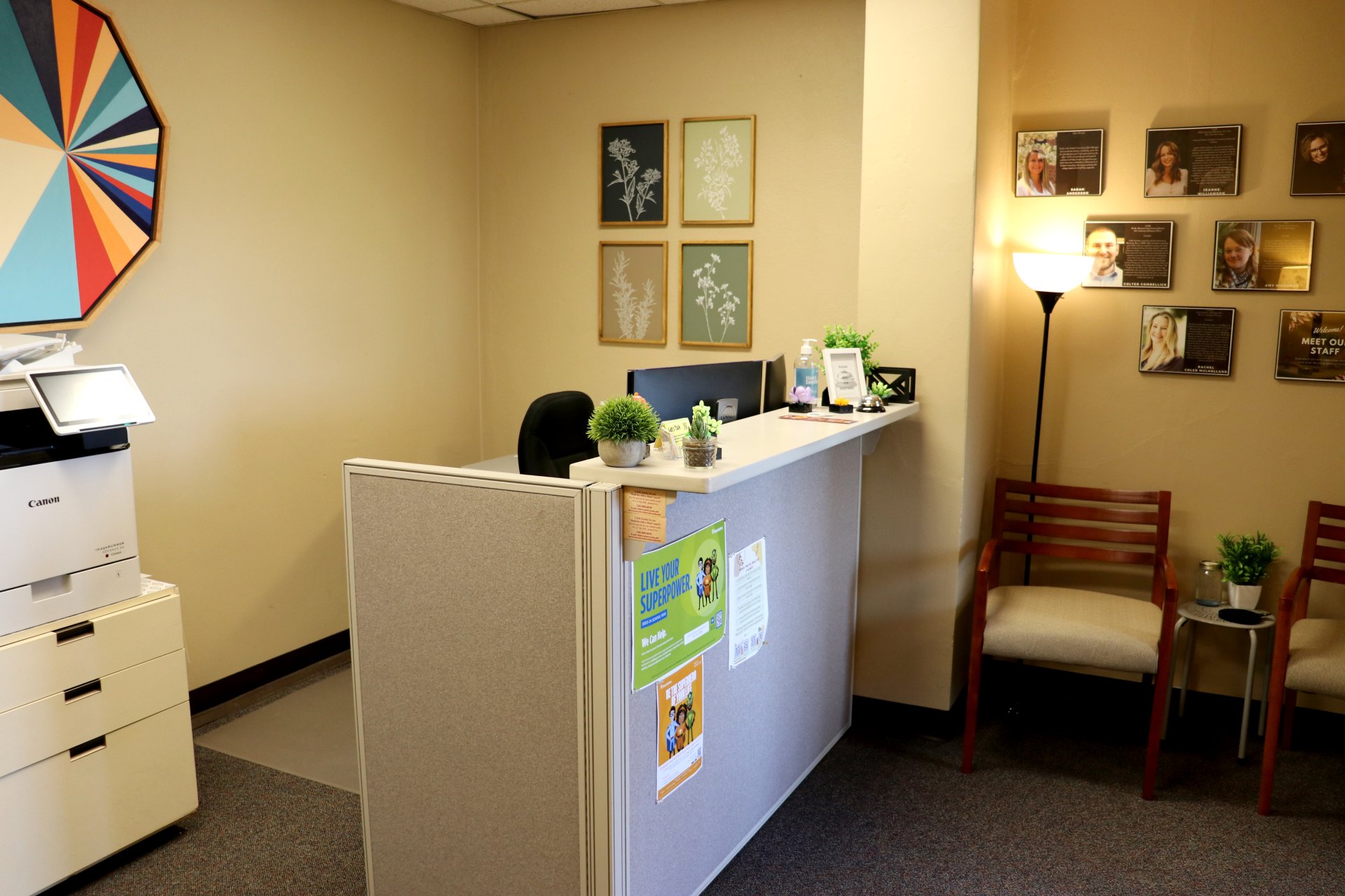 Small, welcoming office space with a front desk. The desk has small plants on top, and behind it is a chair and a wall decorated with framed botanical prints. A nearby wall features staff photos and information, along with two wooden chairs for seating. A floor lamp provides additional lighting, and a printer is positioned on the left side.