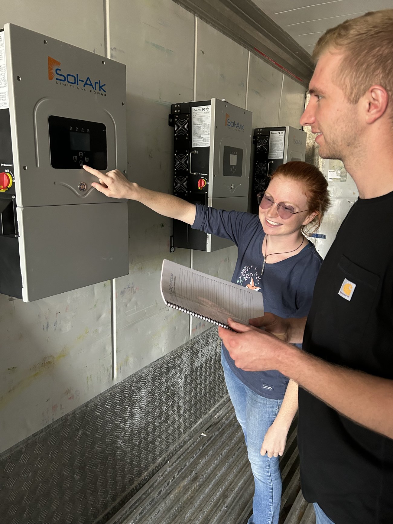 Two people are interacting with a battery system. One person is pointing at the screen of a 'Sol-Ark' unit, while the other holds a notepad and observes. Both are smiling and appear to be in a technical workspace, with multiple battery units mounted on the wall behind them.