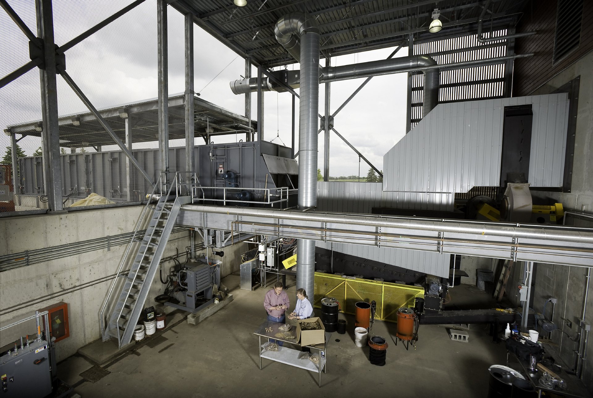 Two individuals are standing at a table inside a large industrial facility. They appear to be examining materials, possibly biomass samples. The space is filled with industrial equipment, including large ducts, machinery, and metal platforms. The area is well-lit, with visible piping and a staircase leading to higher platforms.