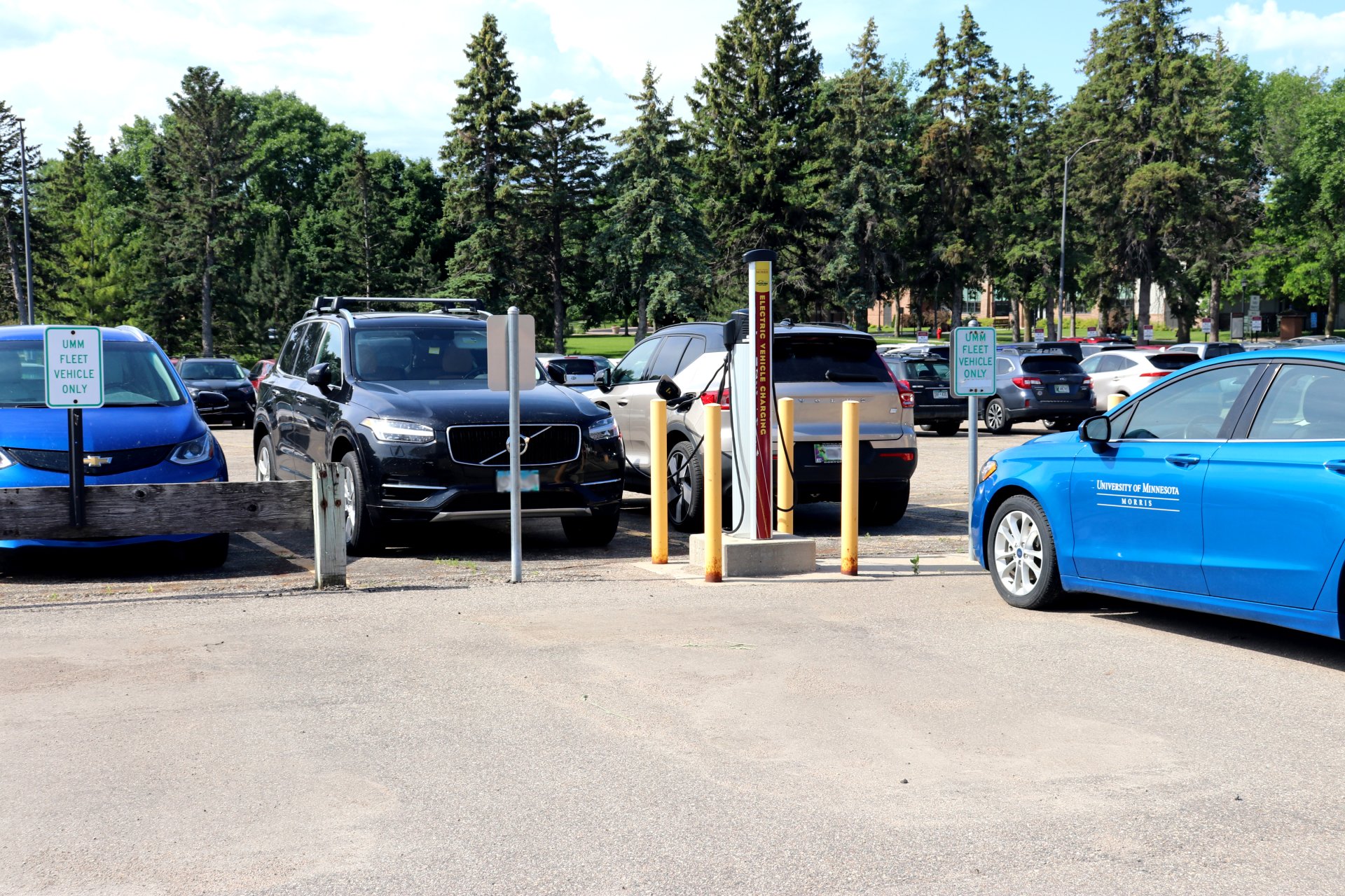 Parking lot with electric vehicle charging station, several cars, and 'UMM Fleet Vehicle Only' signs. Trees visible in the background.