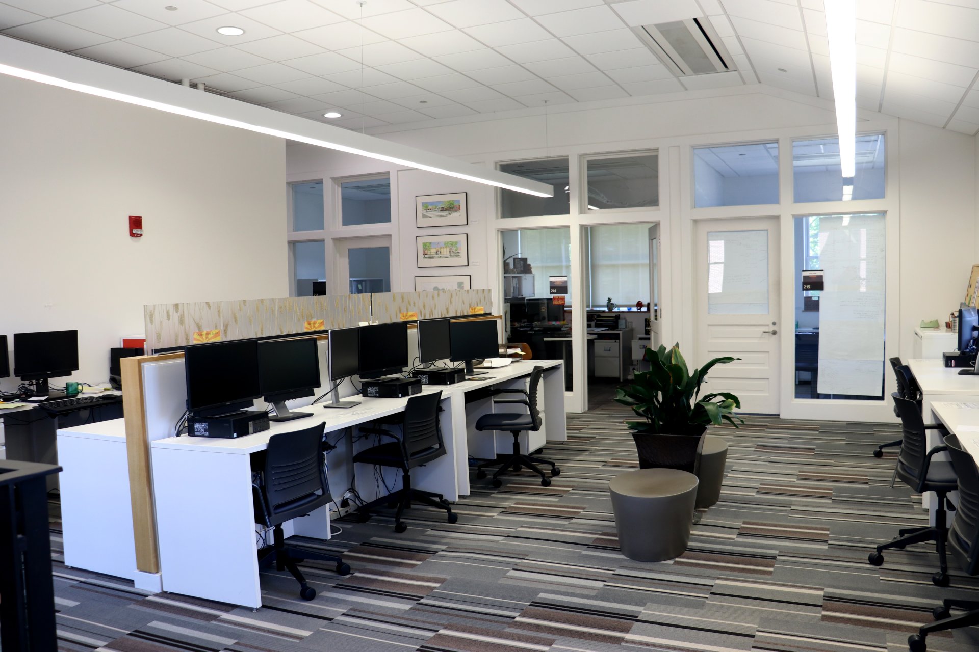 Office space with rows of desks, computer monitors, and chairs. Glass doors lead to private offices in the background. A plant and small stools are placed in the center.