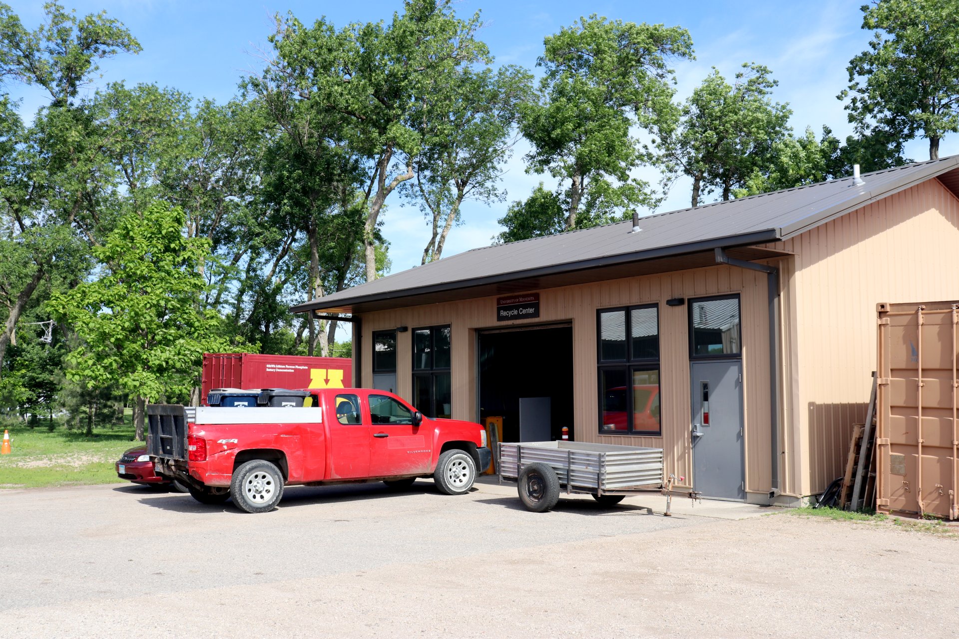 Exterior of a recycling center with a red truck and a trailer parked in front. The building has tan siding, large windows, and a sign above the garage-style door. Trees are in the background.