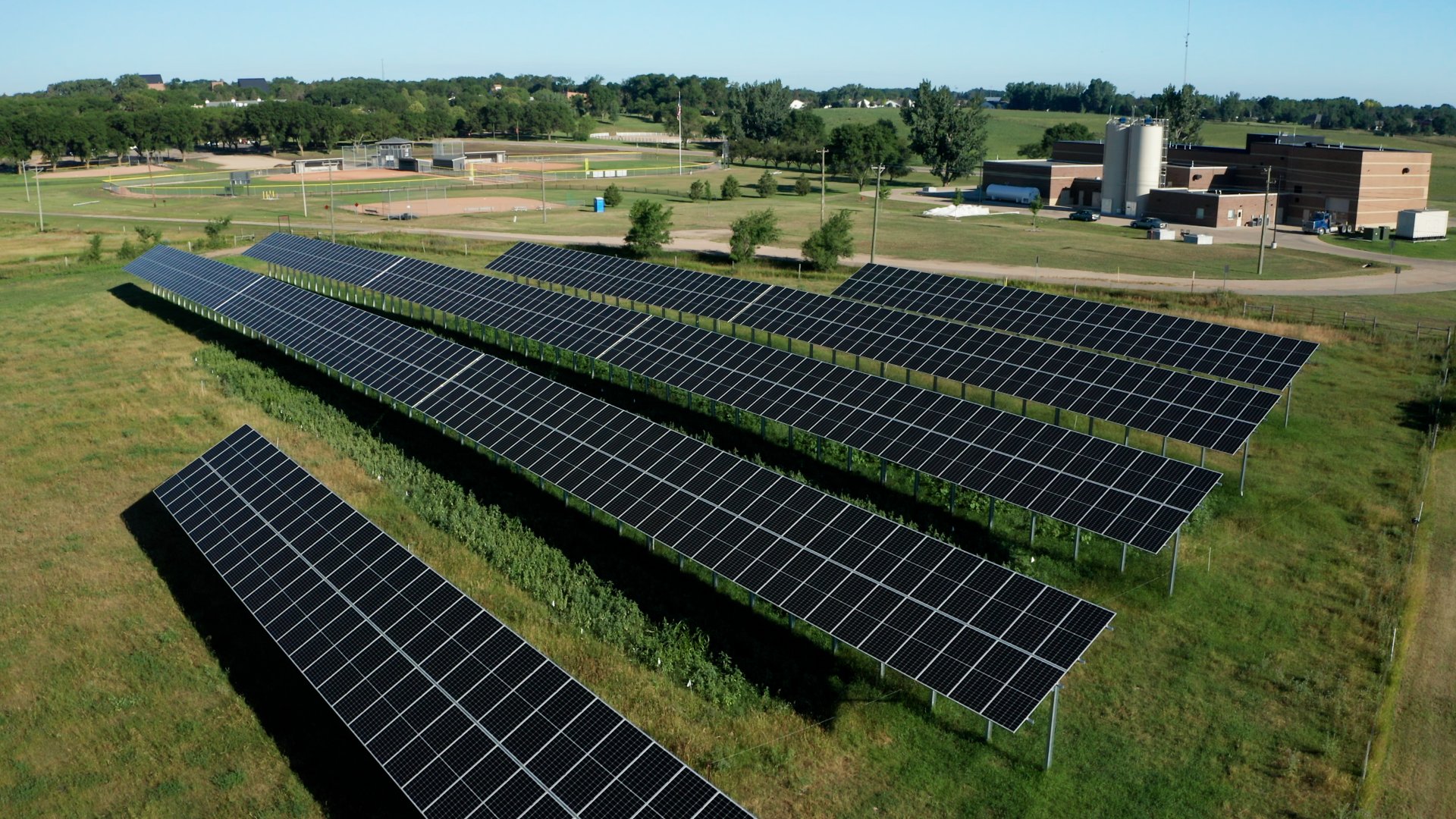 A large array of solar panels in a grassy field, with buildings, a water tower, and sports fields in the background.