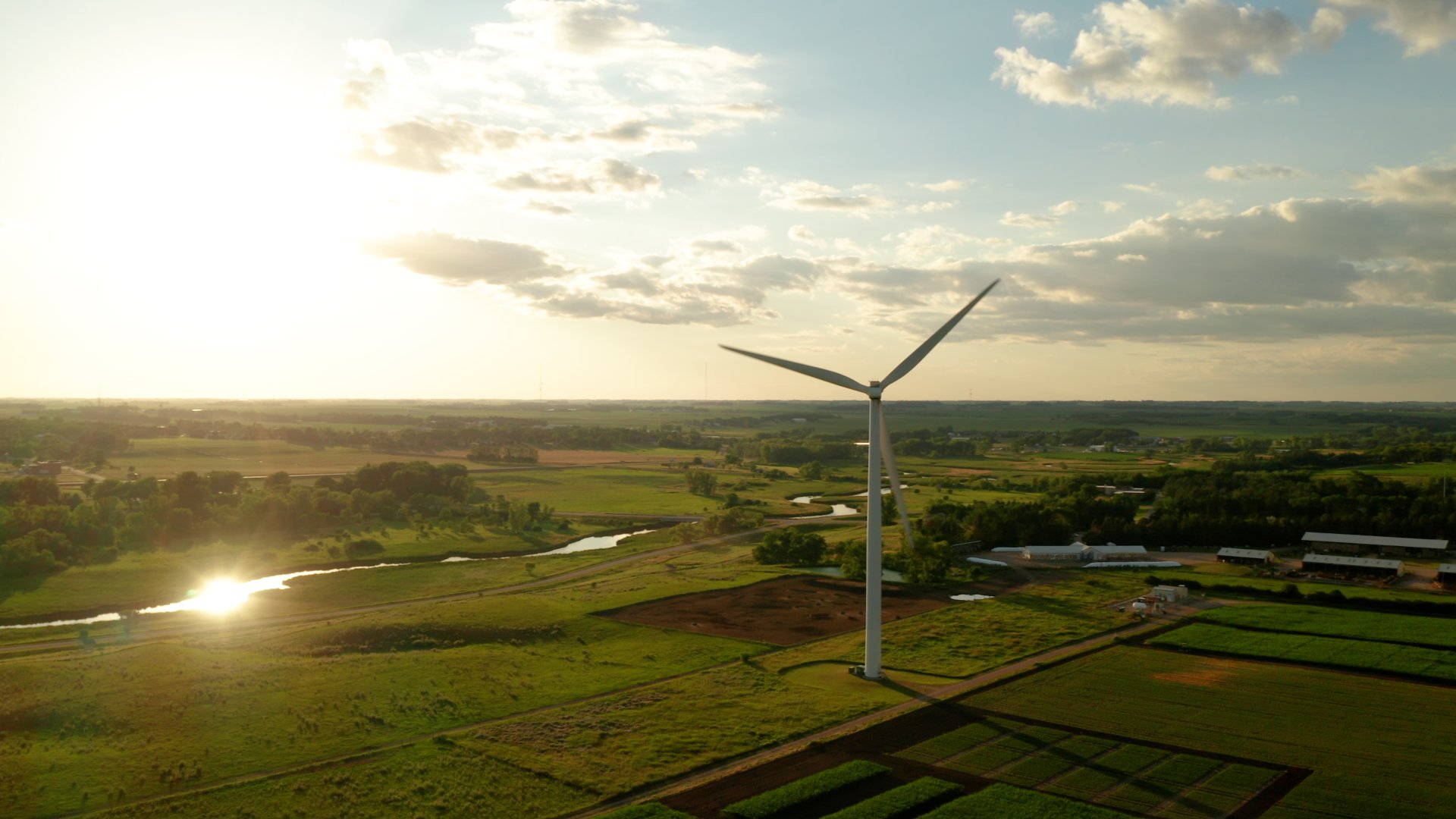 A wind turbine in an open, rural landscape at sunset, with green fields and a reflective body of water nearby.