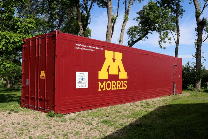 A large red shipping container labeled with the yellow 'M' logo of the University of Minnesota Morris, used for a 90kWh lithium ferrous phosphate battery demonstration. The container sits on a grassy area surrounded by trees under a clear blue sky.