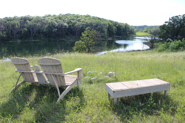 Two wooden Adirondack chairs and a small table are placed on a grassy hillside overlooking a calm lake, surrounded by dense trees and rolling hills under a bright sky.
