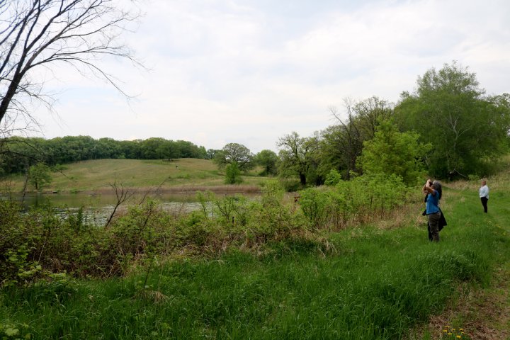 Two people stand along a grassy path near a lake surrounded by trees and bushes. One person is looking through binoculars, while the other stands further back. In the background, rolling hills and more trees can be seen under a cloudy sky.