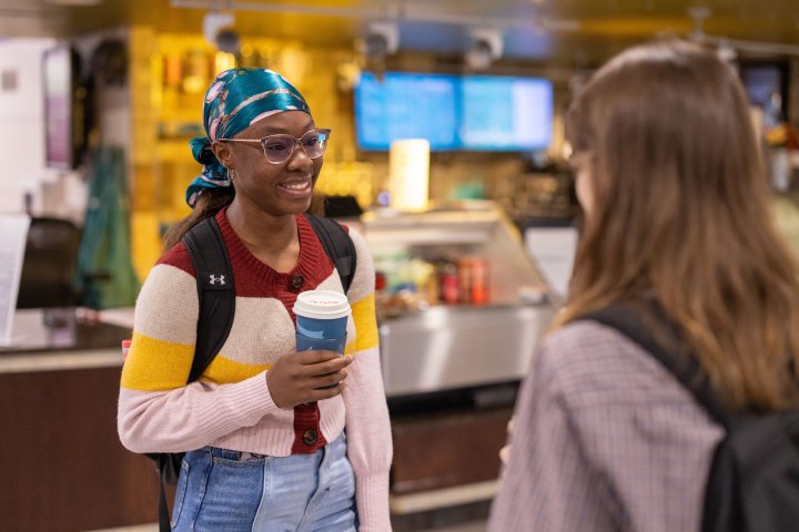 A student wearing a colorful headscarf and a striped sweater holds a coffee cup while chatting with a friend in a café. The café counter with a menu and various drink options is visible in the background, creating a casual and warm atmosphere. The student is smiling, engaged in conversation.