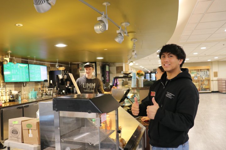A student in a black hoodie gives two thumbs up while standing at the counter of a campus café. Behind the counter, a worker in a hat is smiling. The menu is visible on screens in the background, along with various café items like syrups and cups.