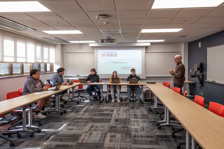 A classroom with a group of students sitting at tables in a U-shaped formation, each working on laptops. A professor stands at the front of the room, speaking to the students, with a projector displaying a presentation on the screen behind him. The room has large windows, bright lighting, and red chairs.