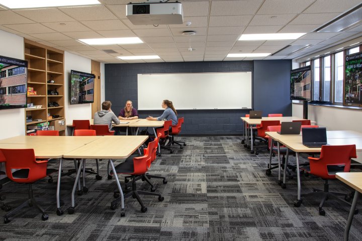 A classroom with three students seated at tables, engaging in a discussion. The room has red chairs, multiple flat-screen displays on the walls, and laptops placed on tables. A large whiteboard is positioned at the front of the room, and there are shelves with games and supplies on the left side.