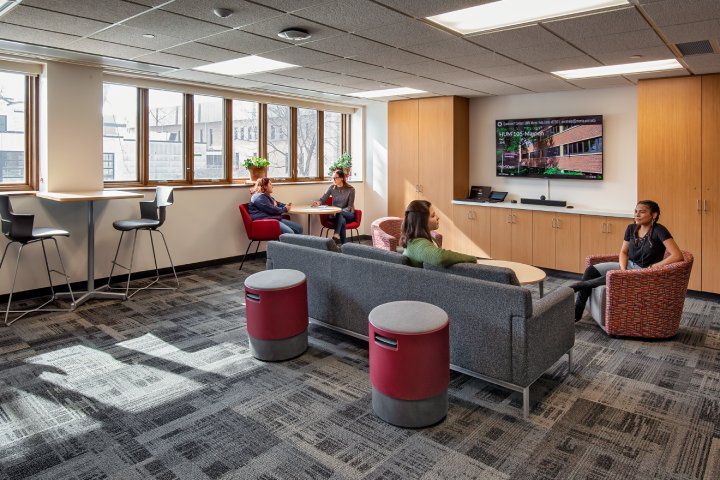 A lounge area with four people seated in comfortable chairs and couches. The room has large windows allowing natural light, a flat-screen display on the wall, and high-top tables with chairs by the windows.