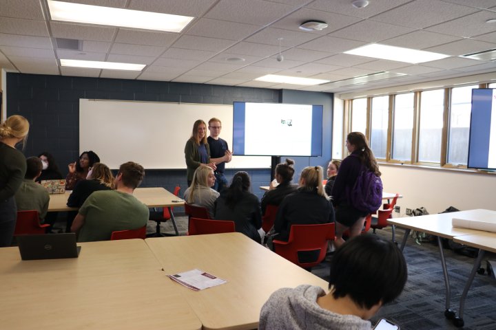 A classroom filled with students sitting at tables. Two students stand in front of the class presenting, while others engage in conversation or listen. Multiple monitors display information behind them, and sunlight streams in through the large windows on the right side of the room.