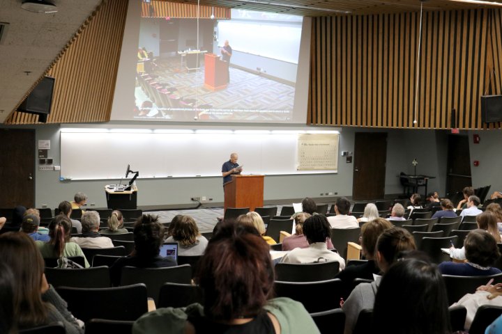 A lecturer speaks at a podium in a large auditorium, addressing an audience of students seated in rows of chairs. A projected screen above the speaker shows a live feed of the presentation. The room has wood-paneled walls and a periodic table of elements is displayed on the wall to the right of the speaker.