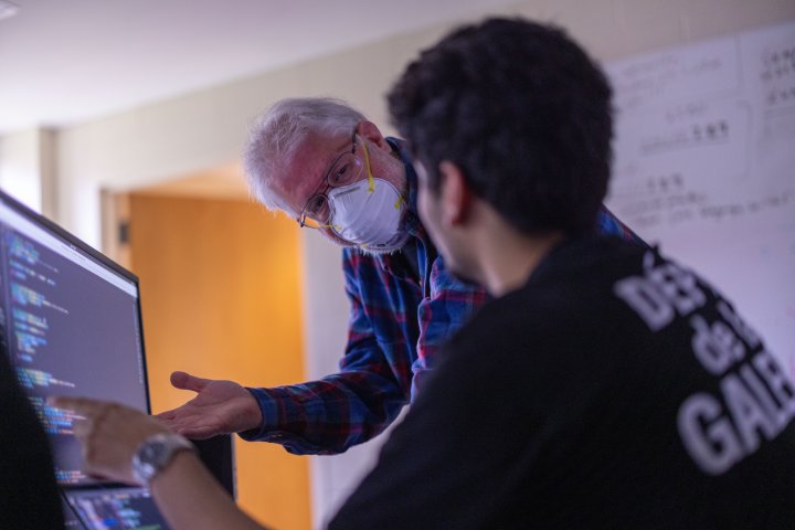 
              An older professor wearing a mask leans over a student's shoulder, gesturing toward a computer screen displaying lines of code. The professor appears to be providing guidance or explaining something, while the student listens attentively. The room has a soft, dim light.