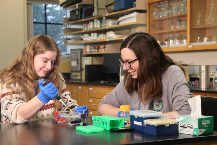 Two women in a science laboratory work together on an experiment. The woman on the left, wearing a sweater and blue gloves, uses a pipette to carefully transfer a liquid into a gel electrophoresis setup, while the woman on the right, wearing glasses and a sweatshirt, watches and smiles.