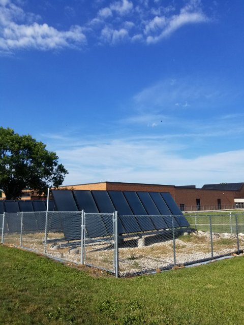 An array of solar panels enclosed by a chain-link fence on a grassy area, with a brick building and a tree in the background beneath a blue sky with scattered clouds.