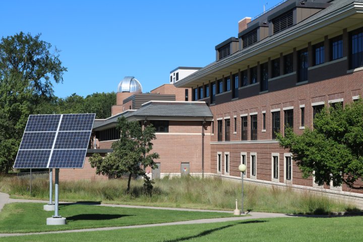 A solar panel mounted on a stand in front of a large brick building with many windows, surrounded by grass and trees. The building features an observatory dome on the roof, and the scene is set under a clear blue sky.